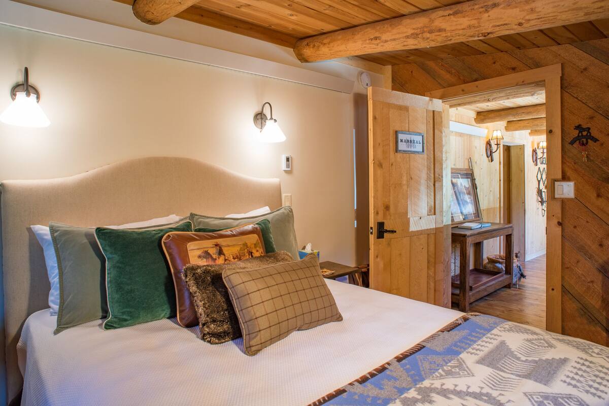 Una cama bien hecha con varias almohadas y una colcha estampada, paredes de madera y una puerta que da a otra habitación.