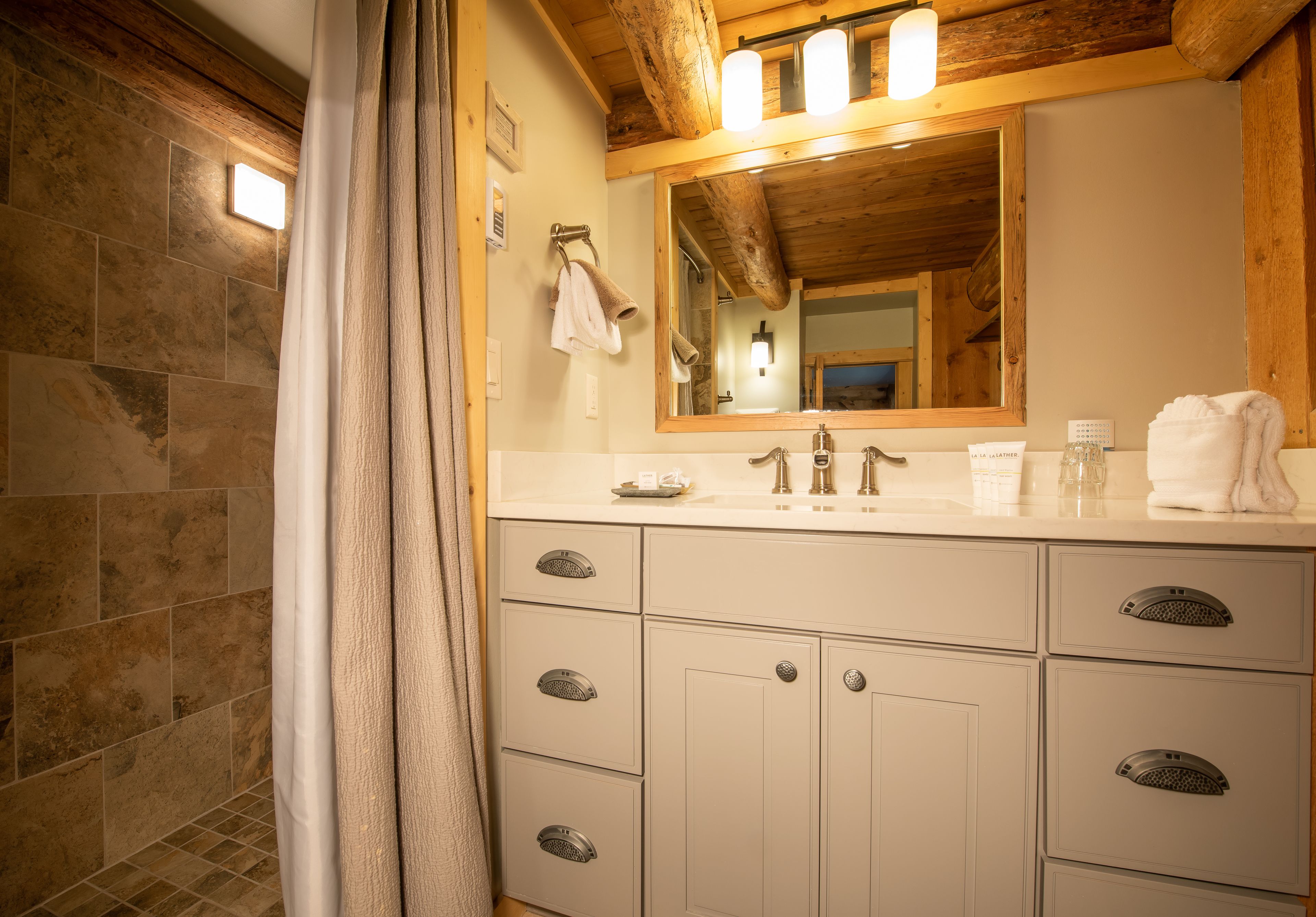 A bathroom with a stone-tiled shower area, a wooden ceiling, and a large mirror above a white sink with a towel hanging on the wall.