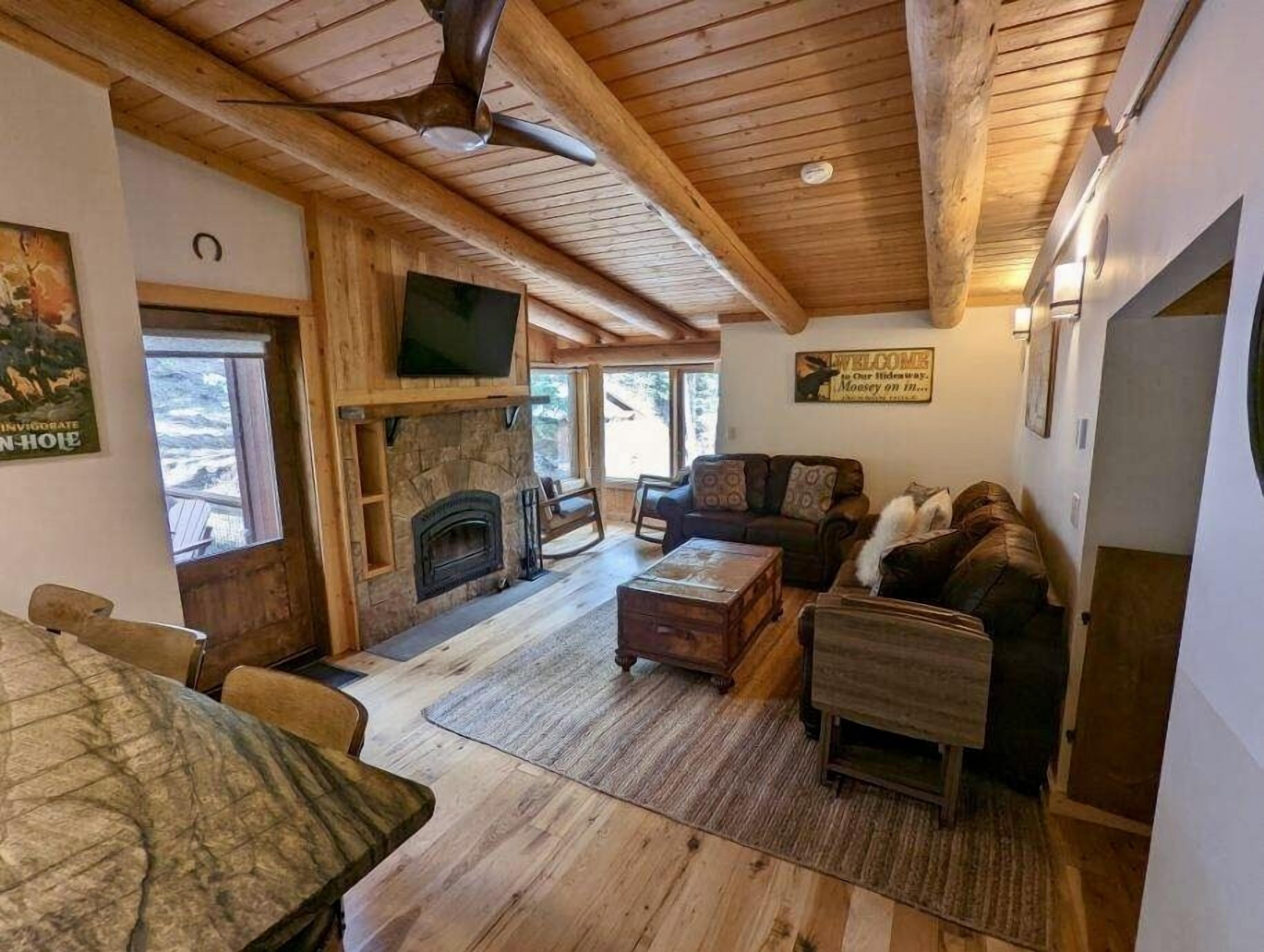 A cozy cabin interior with a stone fireplace, wooden ceiling beams, a television, and a couch.