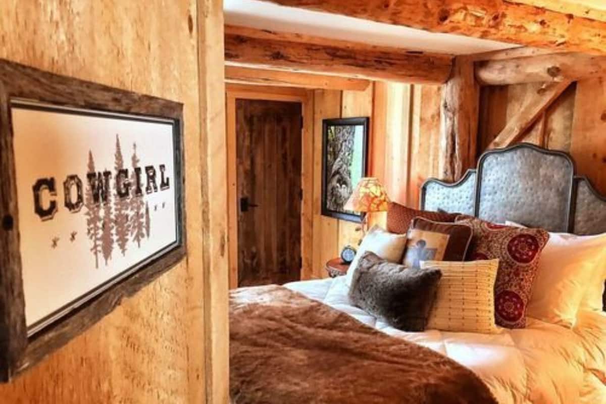 Un dormitorio rústico de madera con un letrero enmarcado que dice 'COWGIRL', una cama con muchos almohadones y una mesita de noche con una lámpara.