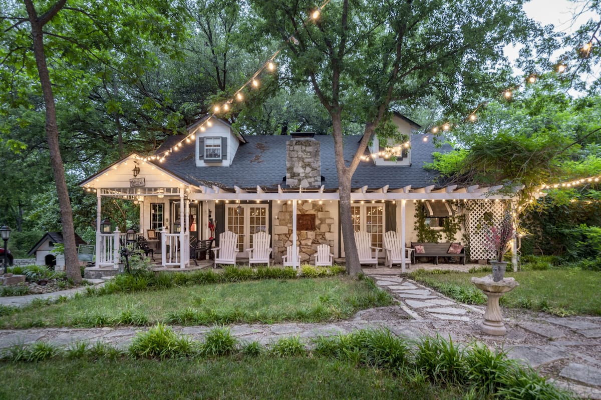 A cozy cottage with a stone chimney, a porch with white chairs, and string lights.