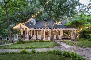 A cozy cottage with a stone chimney, a porch with white chairs, and string lights. - Country Woods Inn (Glen Rose)