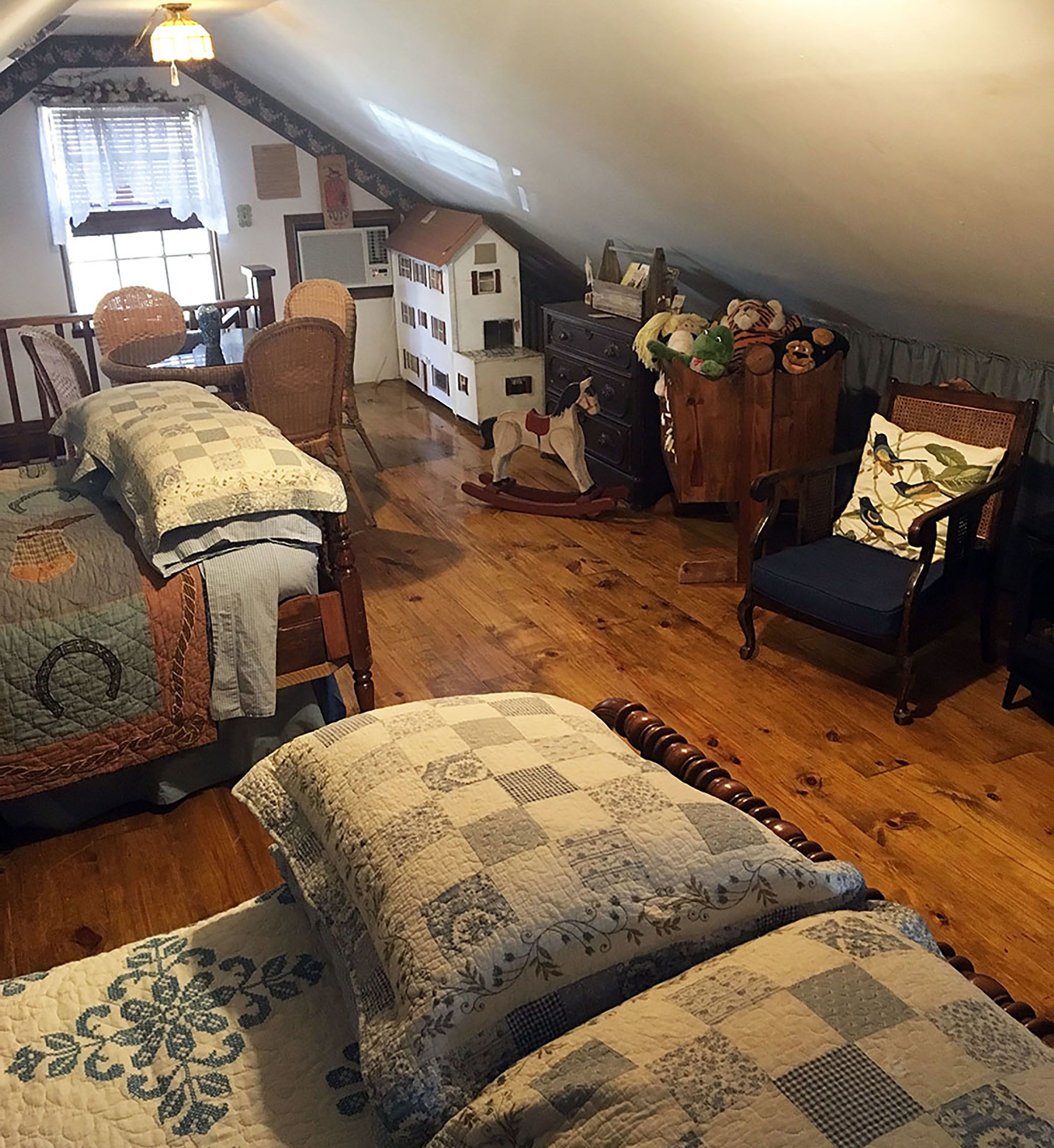 Une chambre confortable située dans les combles, avec un plancher en bois, un lit recouvert d’une courtepointe, un fauteuil à bascule et une commode ornée de peluches.