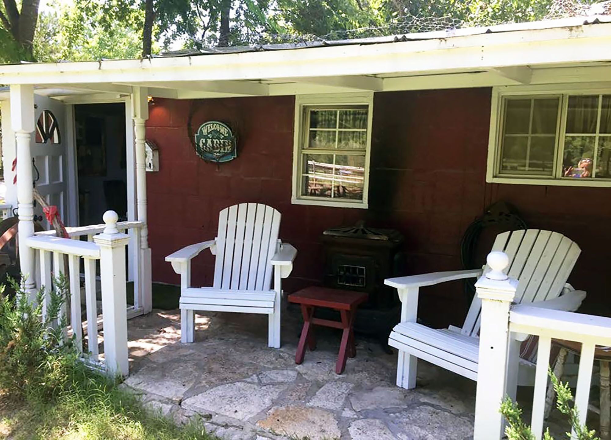 Un porche abrité avec deux fauteuils Adirondack et une petite table, un sol en pierre et une pancarte de bienvenue accrochée au mur.
