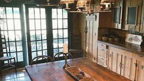 A wooden dining table with a long wooden bench, a candle, and a tray with pinecones and nuts. There are hanging lamps with animal silhouettes, wooden cabinets, and a window with a view of the outdoors. - Country Woods Inn (Glen Rose)