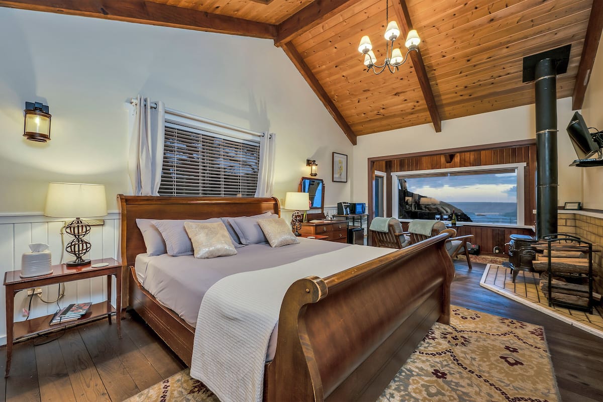 A bedroom with a wooden bed, a nightstand, a lamp, a window with blinds, a view of the ocean, a fireplace, and a desk with a computer.