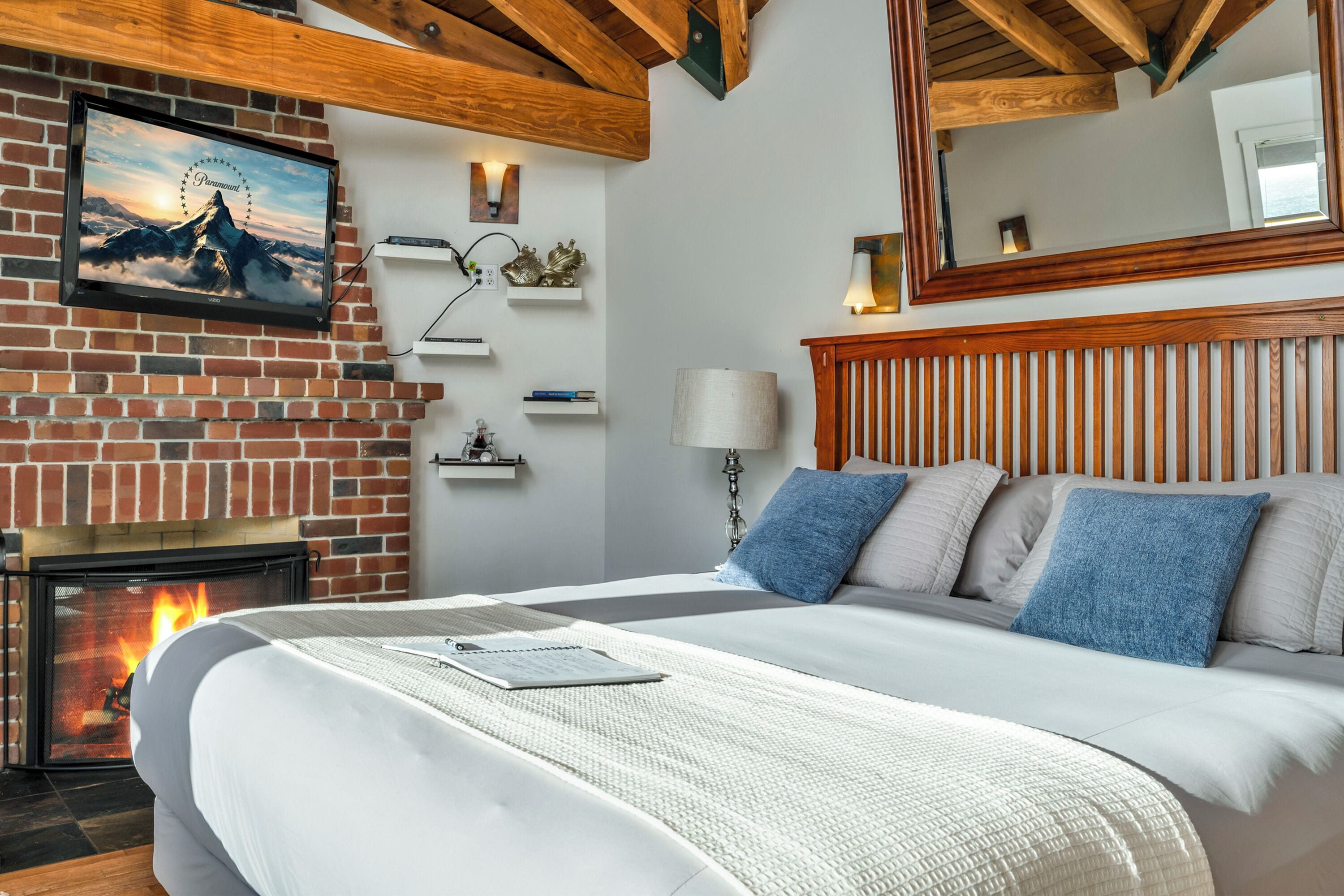 Un dormitorio con chimenea de ladrillo, cama con ropa de cama blanca, televisor montado y viga de madera en el techo.