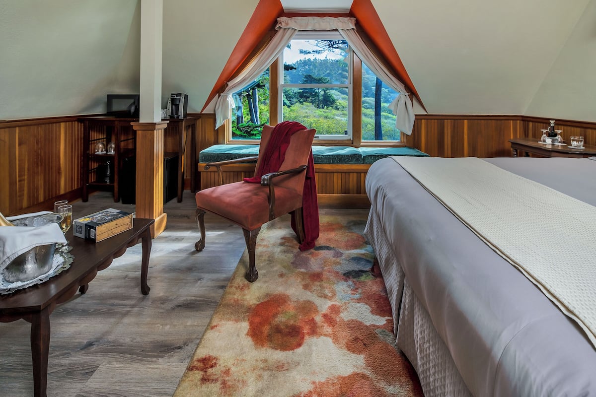 A bedroom with a large window, a bed, a chair, a table with a bowl and a box, and a wooden cabinet.