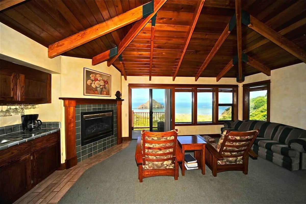 A cozy living area with a fireplace, wooden ceiling beams, and a view of the ocean.