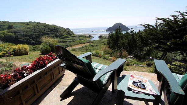 Une terrasse avec deux chaises vertes et une table, d’où l’on peut admirer un paysage côtier avec des collines et l’océan.