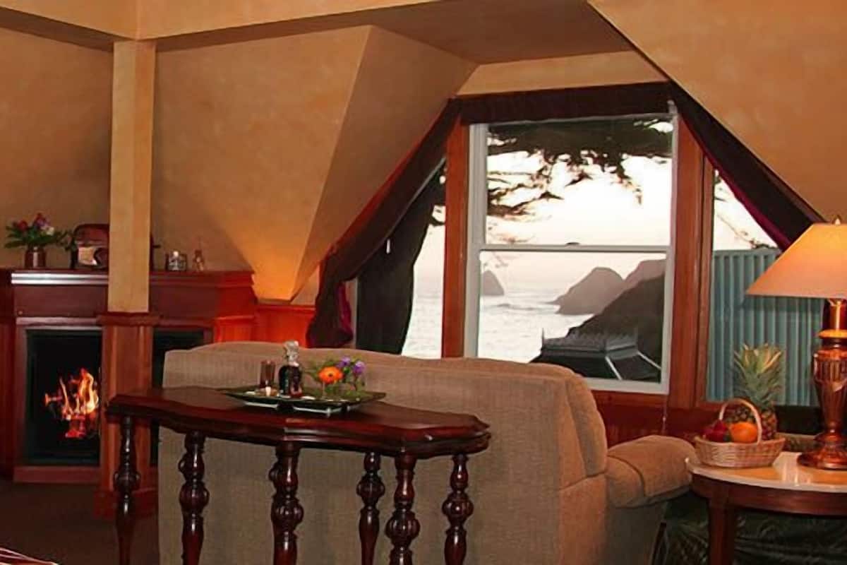 A room with a bed, a fireplace, a table with a lamp, and a view of the ocean through large windows.