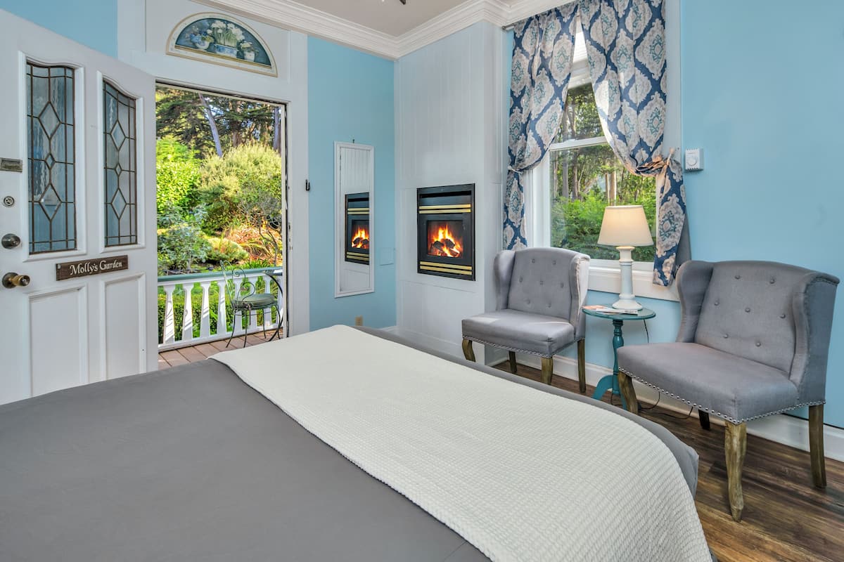 A bedroom with a large bed, two chairs, a small table, a fireplace, and a view of a garden through a window.