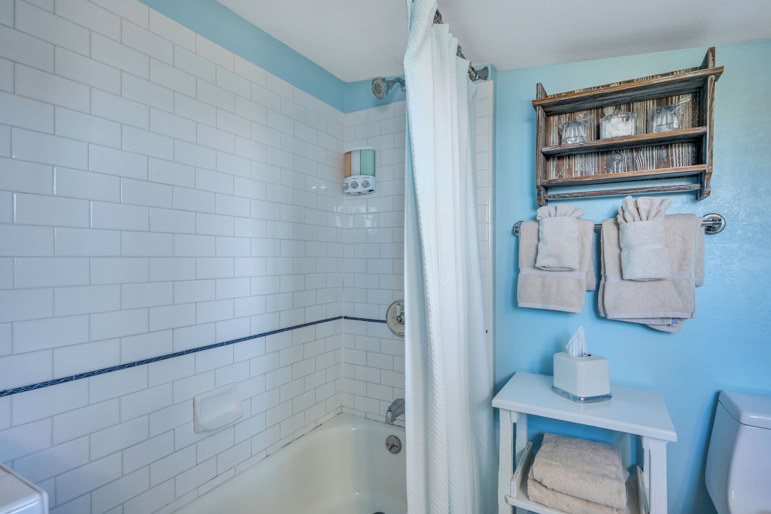 Uma casa de banho com azulejos brancos tipo metrô, cortina de chuveiro, porta-toalhas com toalhas e uma prateleira com frascos.