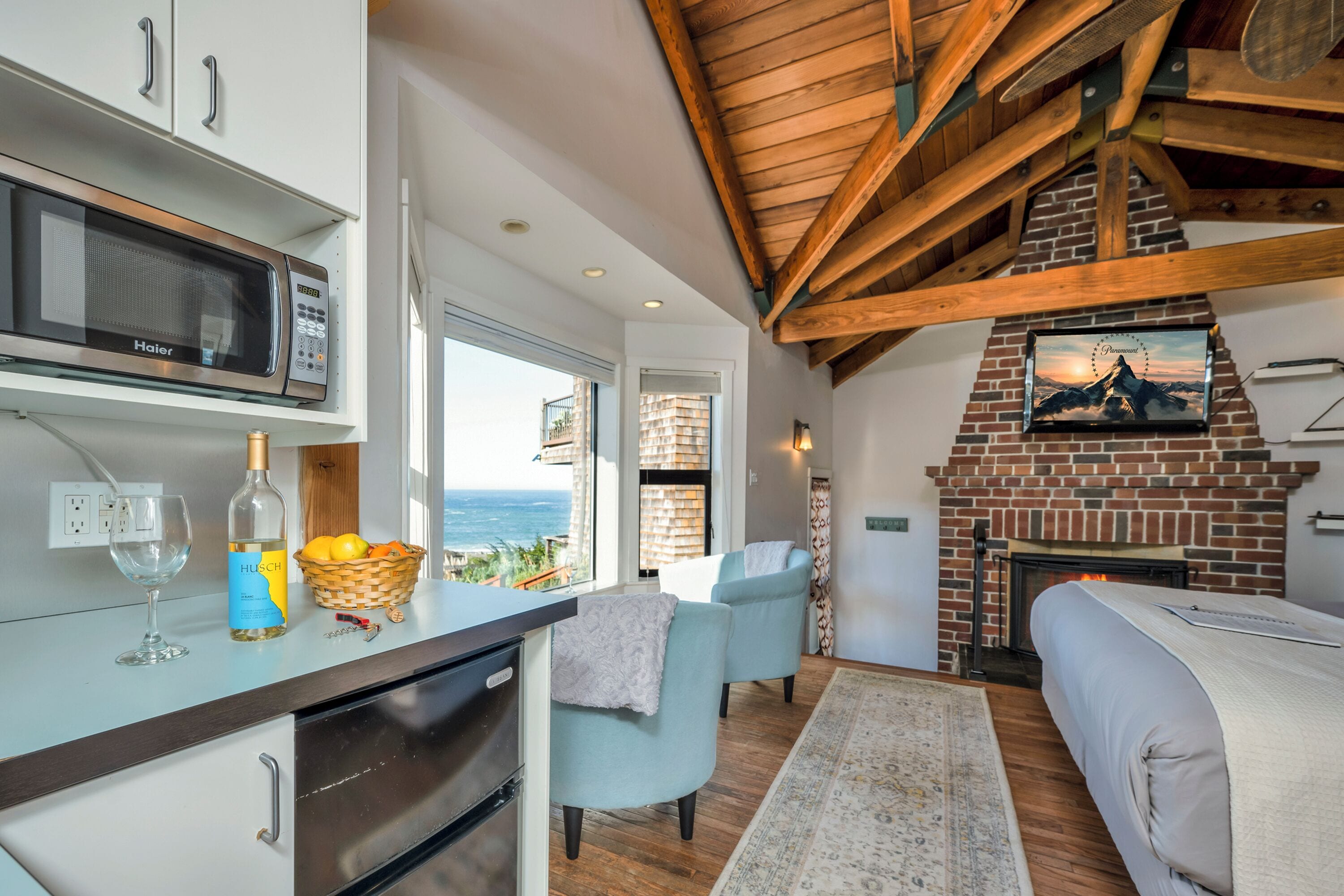 A modern kitchen with a Haier microwave, a wine bottle, and a basket of fruit. There is a fireplace with a brick wall and a painting above it.