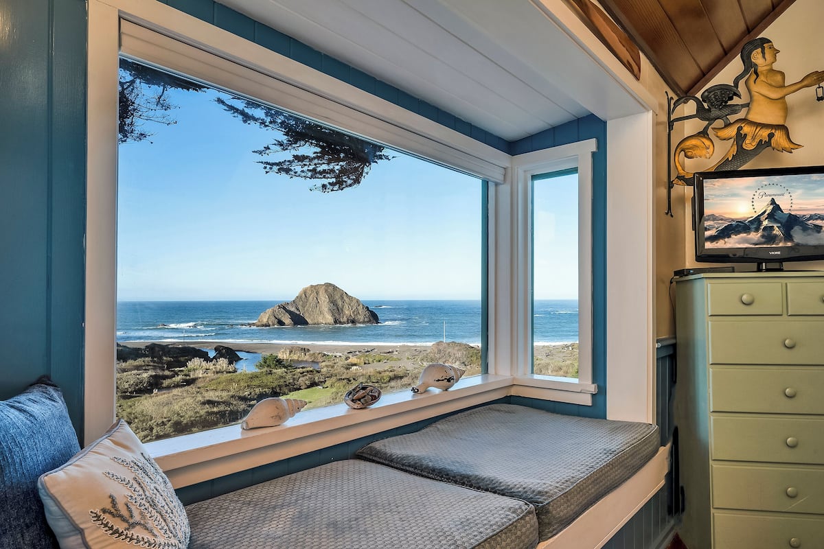 A room with a large window overlooking a beach, a bench, and a dresser with a decorative item.