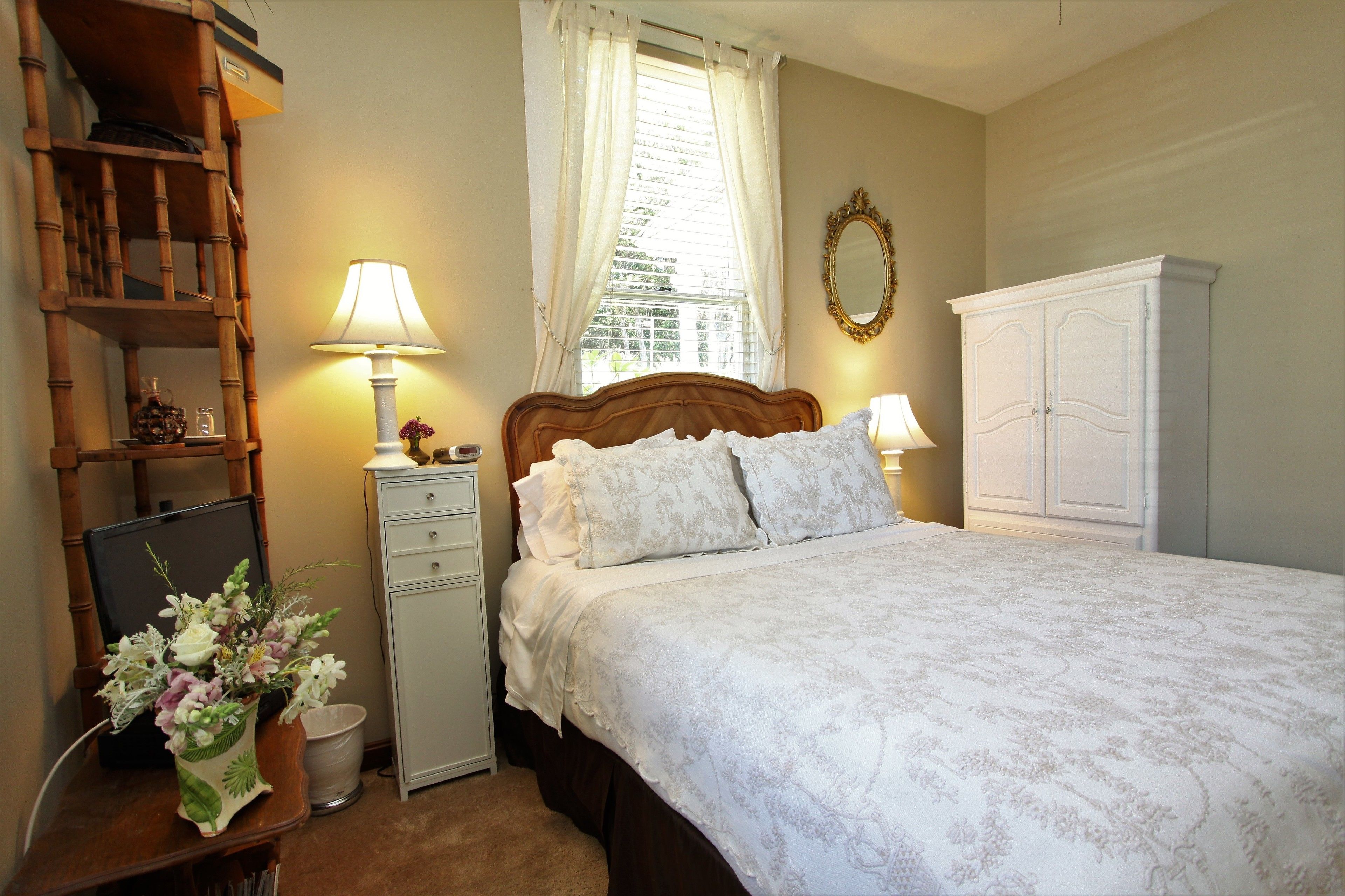 A bedroom with a wooden bed, white bedding, a nightstand with a lamp, a small desk with a computer and a vase of flowers, and a wardrobe.