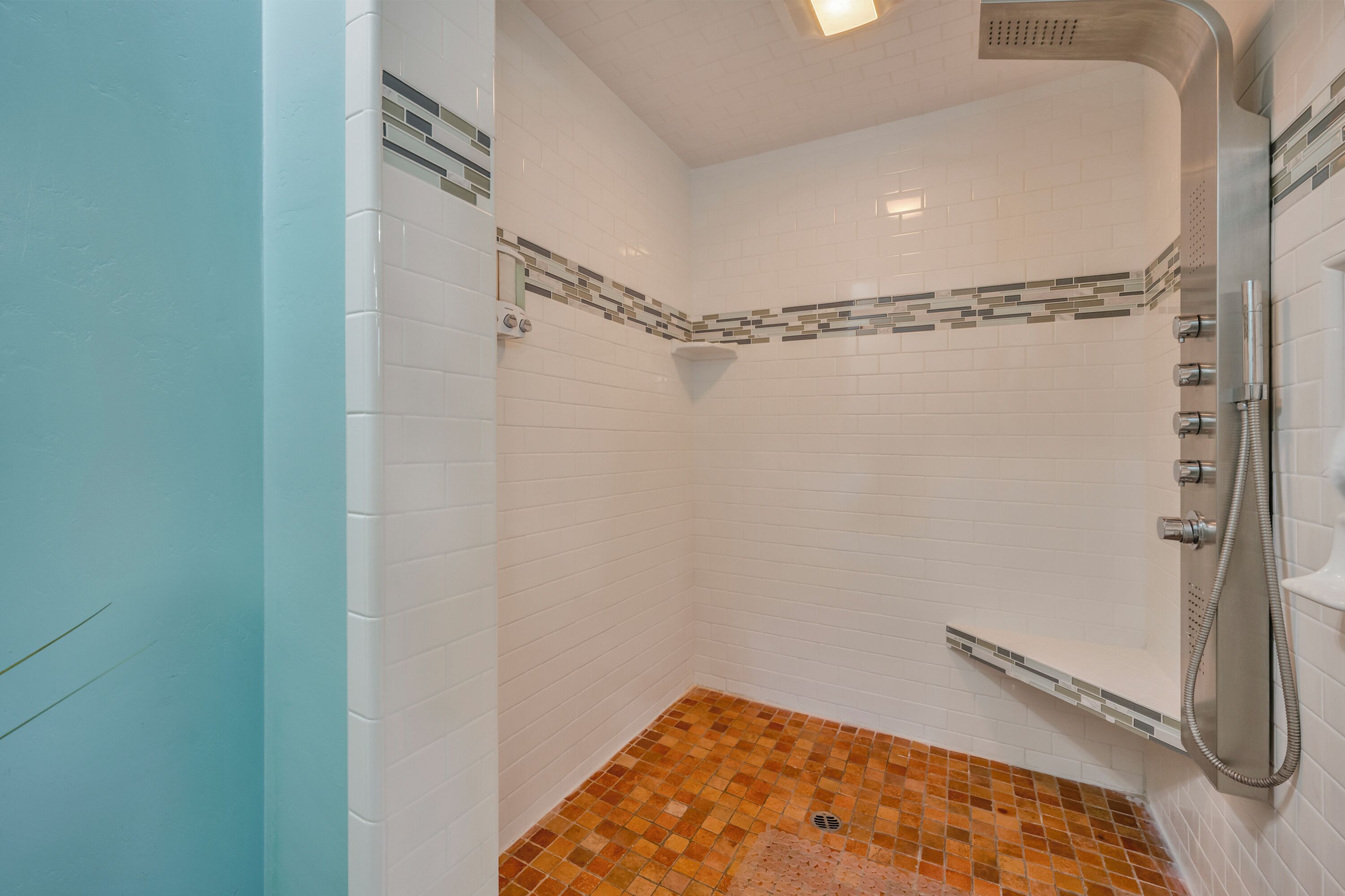 A shower area with white tiled walls, a corner showerhead, and a tiled floor with a mix of orange and brown tiles.