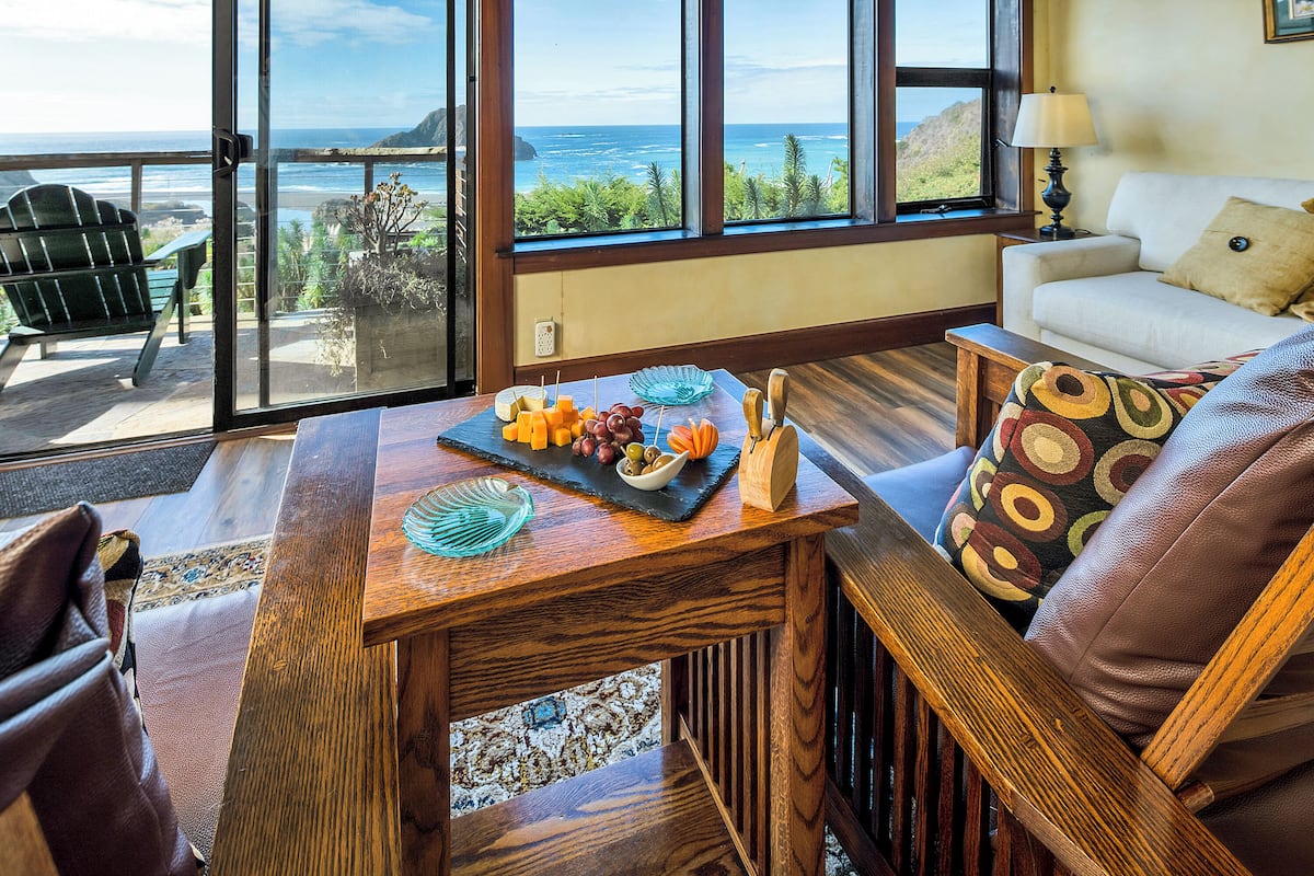 A room with a view of the ocean, a wooden table with fruit and cheese, a green chair, and a white sofa with patterned cushions.