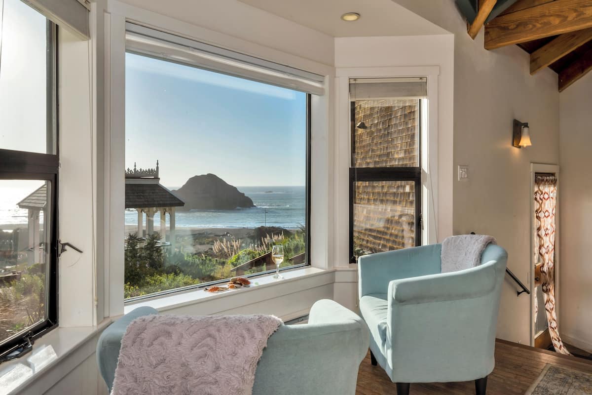 A room with a large window offering a view of the ocean and a distant island. There are two light blue chairs and a fluffy pink throw blanket on one of them.