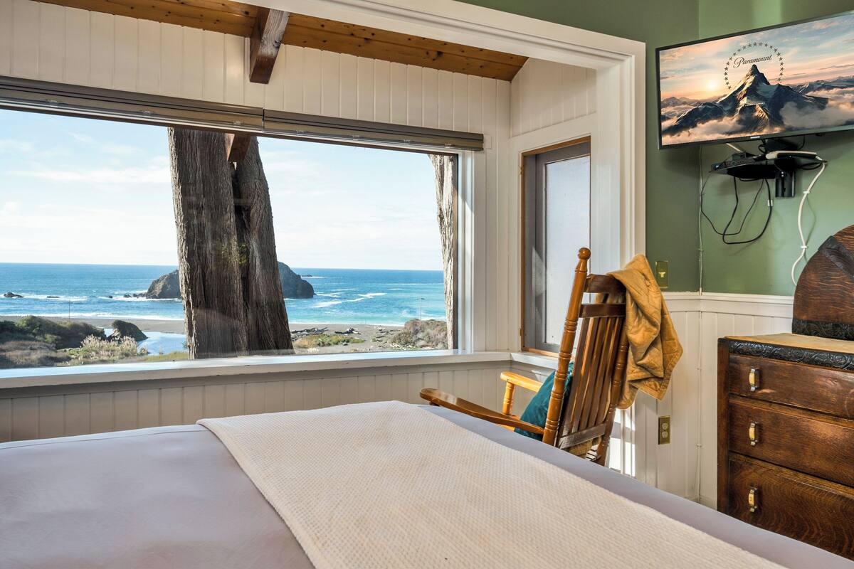A bedroom with a large window overlooking the ocean, a bed, a wooden chair, and a dresser.