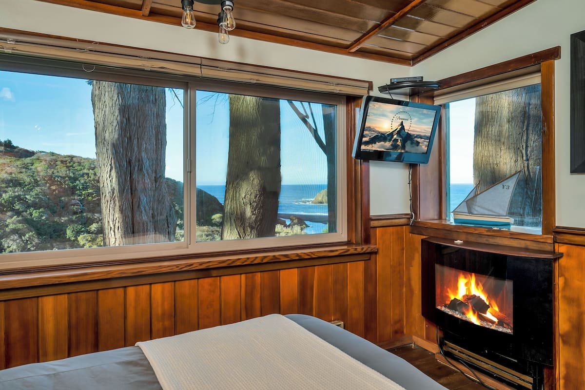 A room with a large window overlooking a scenic landscape, a fireplace, and a television mounted on the wall.