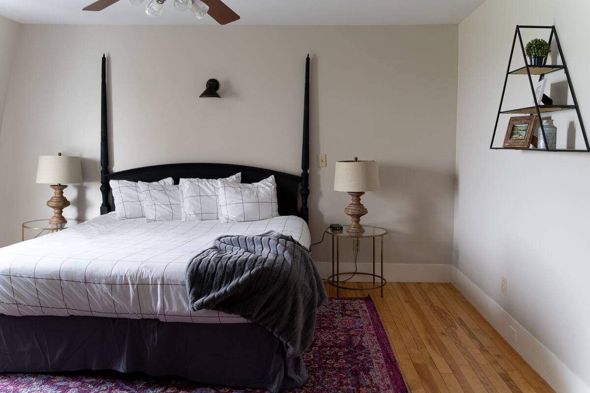 A bedroom with a four-poster bed, two bedside lamps, a small wall-mounted shelf with decorative items, and a wooden floor.