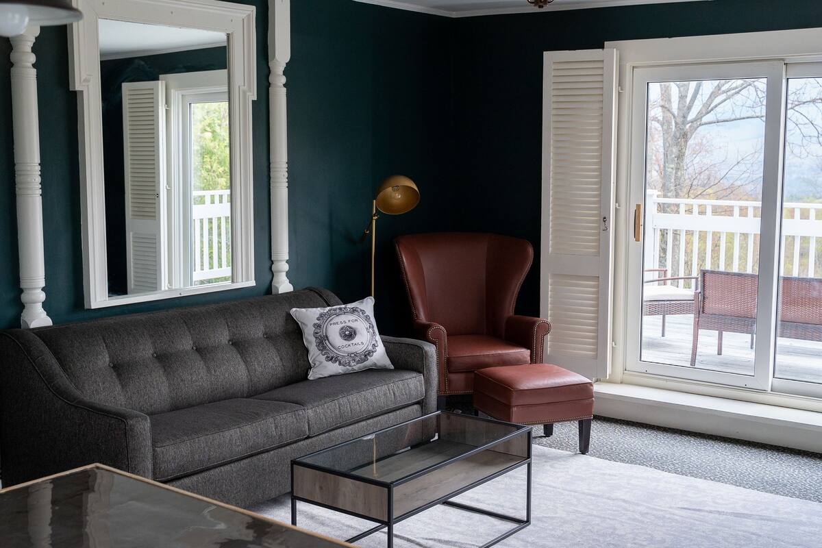 A living room with a dark green wall, a grey sofa with a decorative pillow, a brown leather armchair, a glass coffee table, and a large window with a view of a deck and trees.
