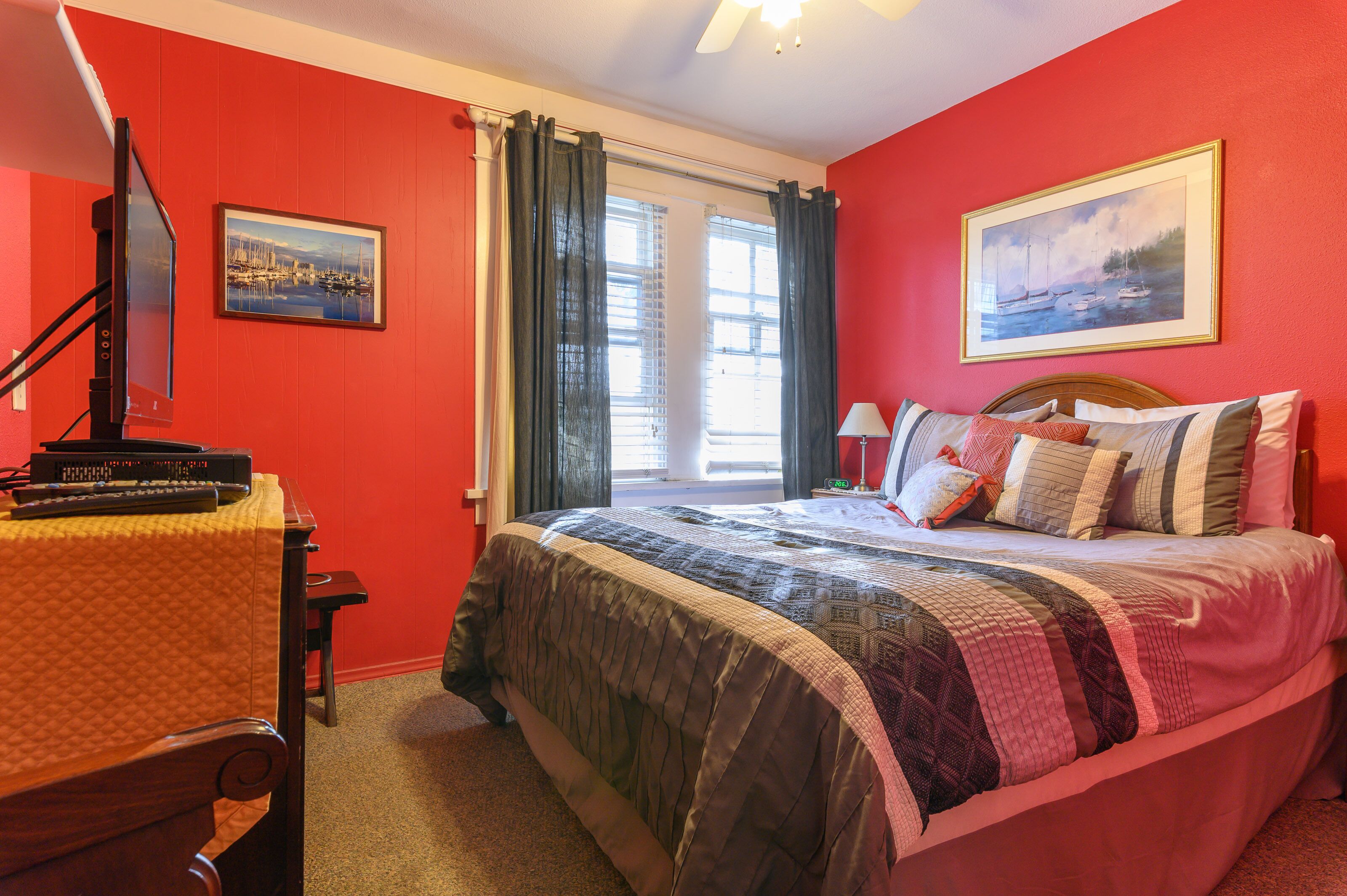 A bedroom with a bed, a desk with a computer, a window with curtains, and two framed pictures on the wall.