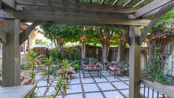 Relax in a serene outdoor space surrounded by lush greenery and potted plants, with a sofa, table, and chairs under a covered pergola.