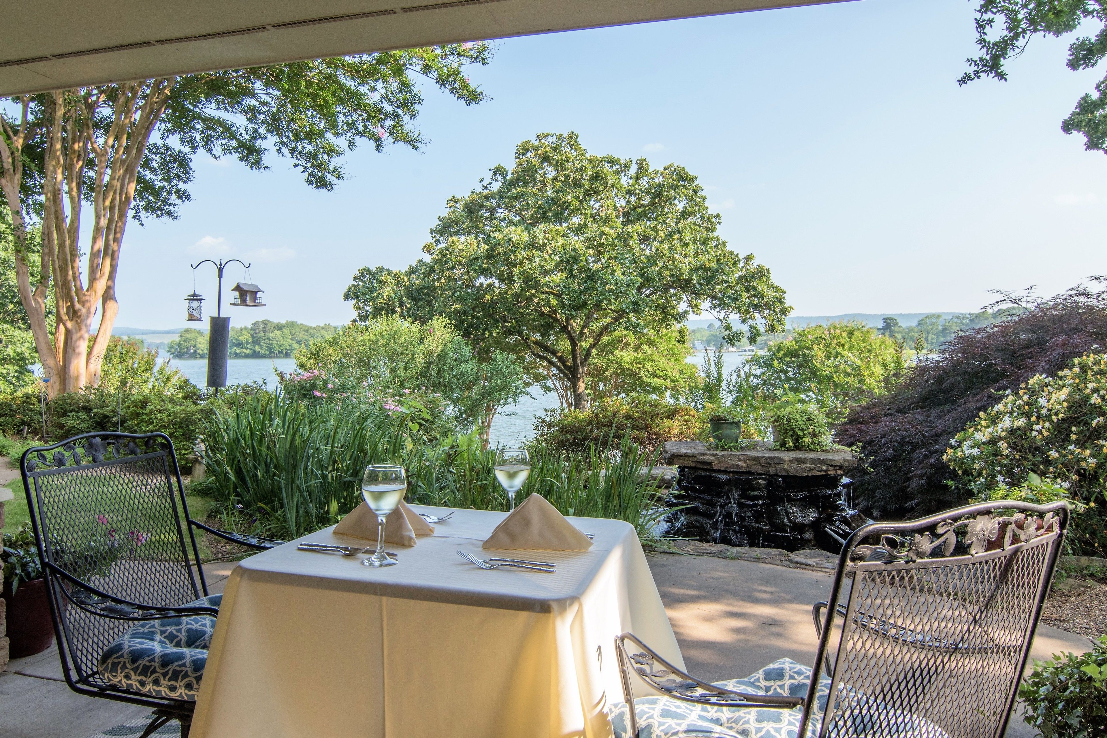 An outdoor dining area with a table set for two, surrounded by chairs, overlooking a lake with trees and a waterfall.
