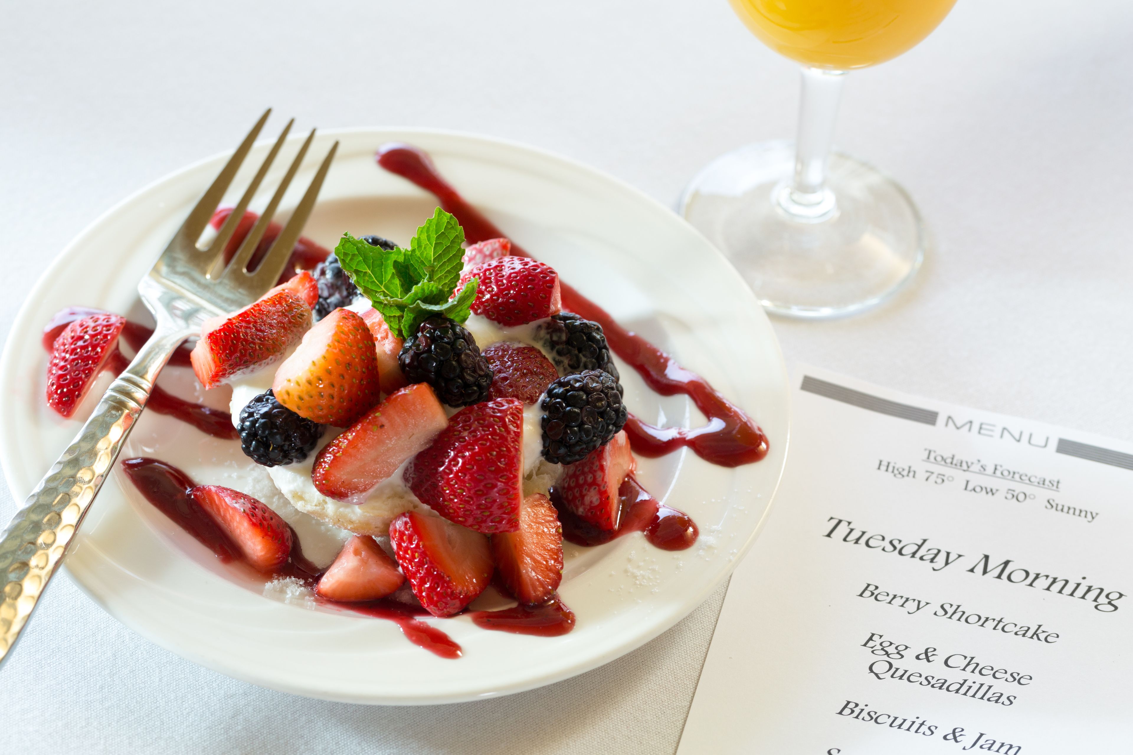 Ein Frühstücksteller mit Erdbeeren, Brombeeren und einer Minzdeko, begleitet von einer Speisekarte mit Angeboten wie Beeren-Shortcake und Quesadillas.