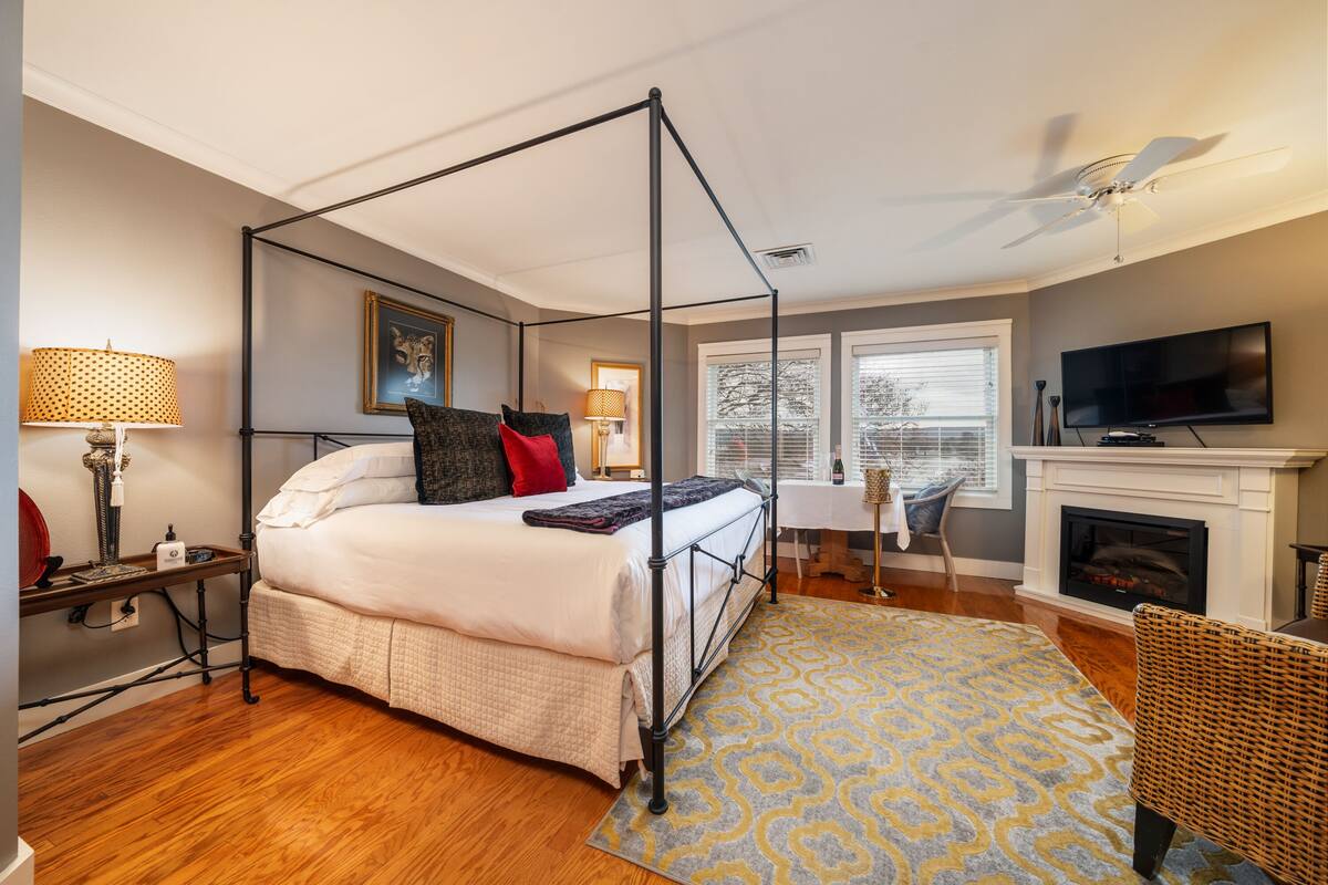 A four-poster bed with a white comforter and red pillows, a fireplace, a television, and a large window with blinds.