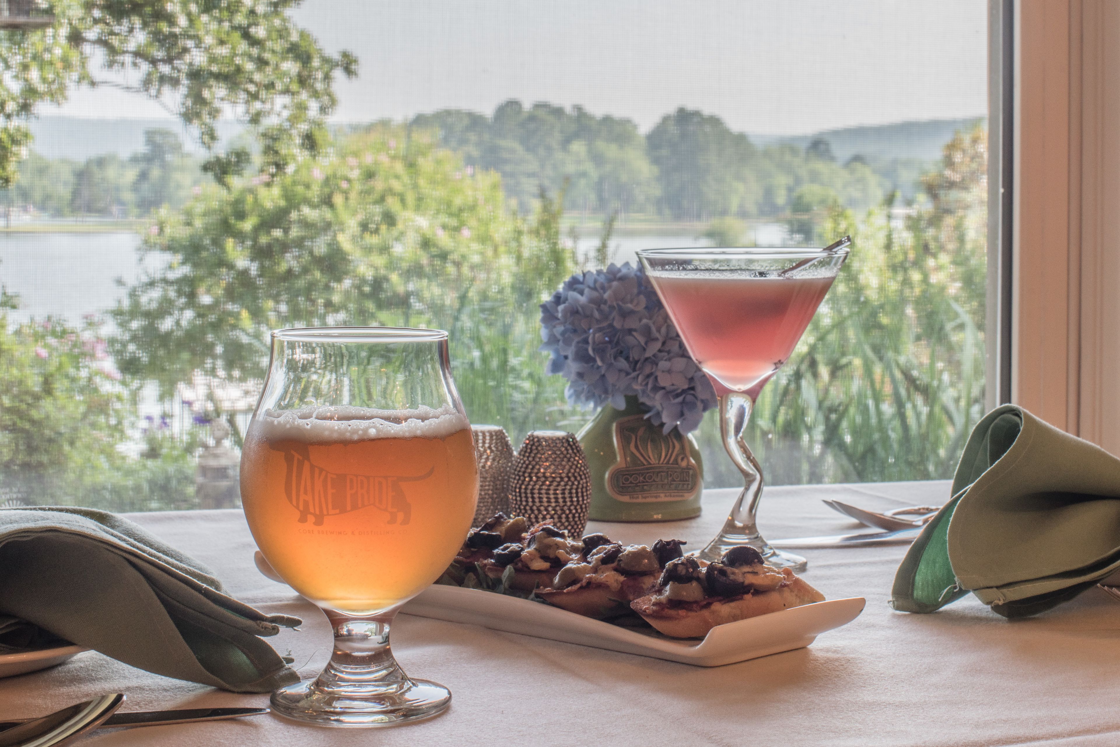 Una mesa con dos vasos de cerveza y un martini, un plato de frutos secos y un jarrón de flores, con una vista panorámica de un lago y árboles al fondo.