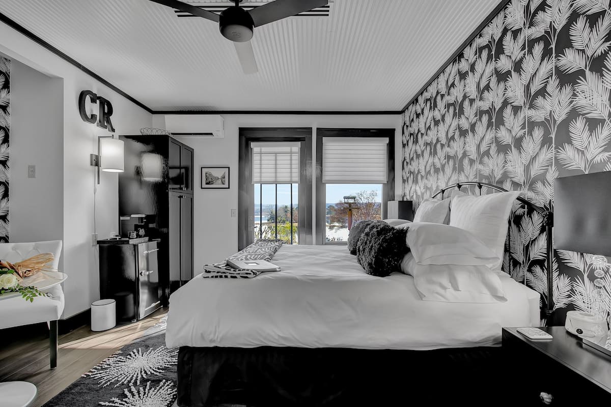 A modern bedroom with a large bed, a ceiling fan, a desk, and a view of the outdoors through large windows.