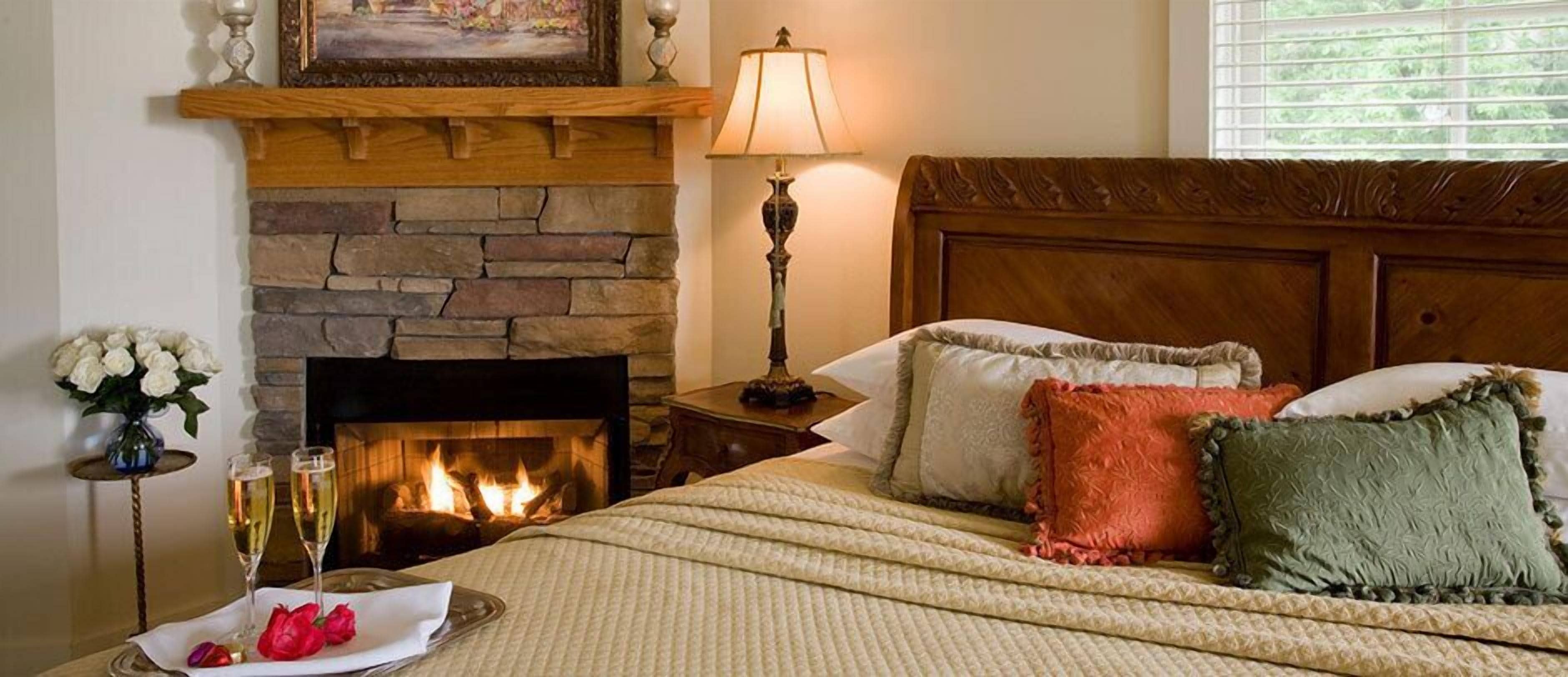 A bedroom with a stone fireplace, a bedside table with a lamp, a bed with multiple pillows, and a vase of flowers on the nightstand.