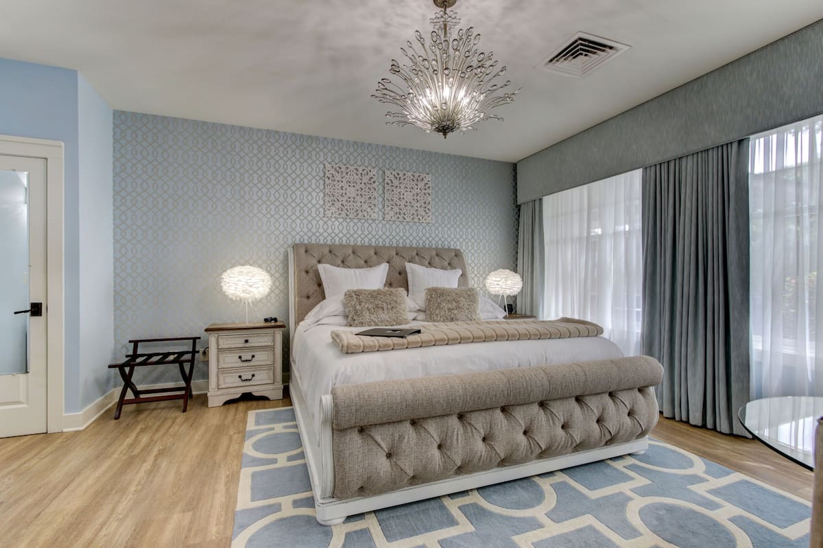A bedroom with a large bed, bedside tables, a chandelier, and a view of the outdoors through large windows.