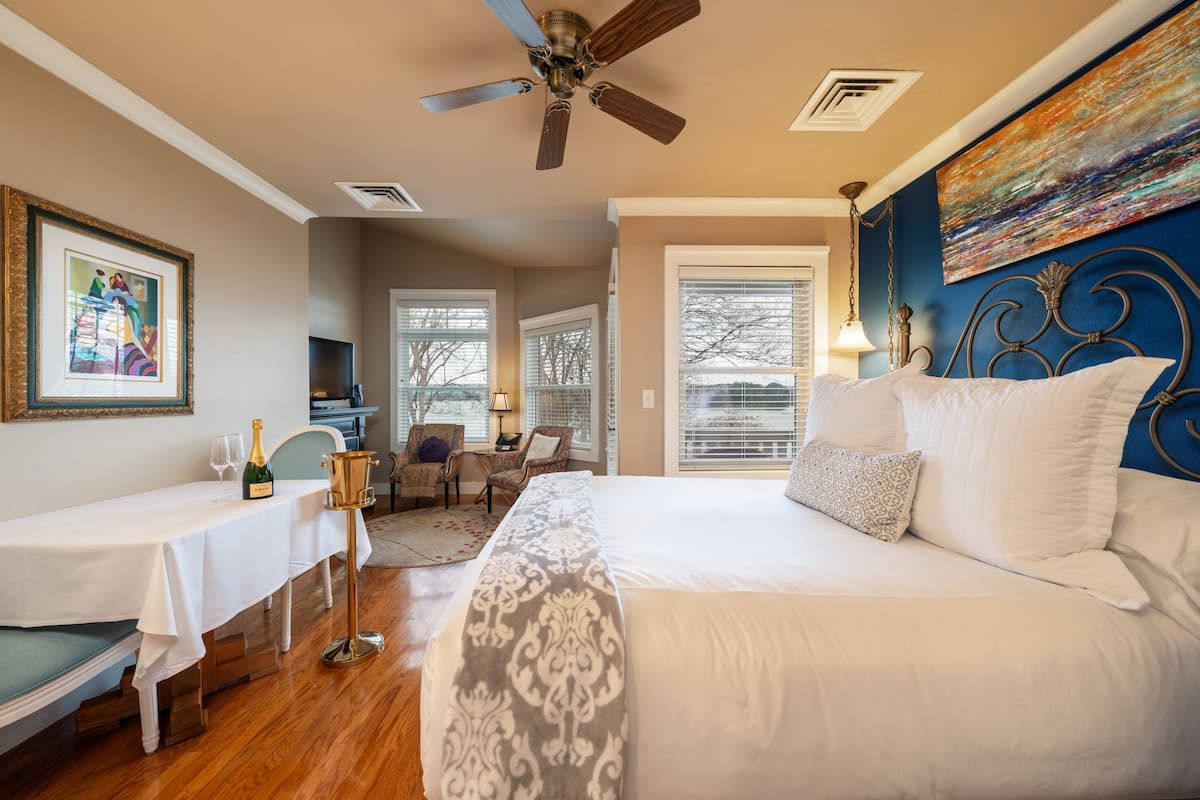 A bedroom with a large bed, a ceiling fan, a painting on the wall, and a dining area with a table set for two.