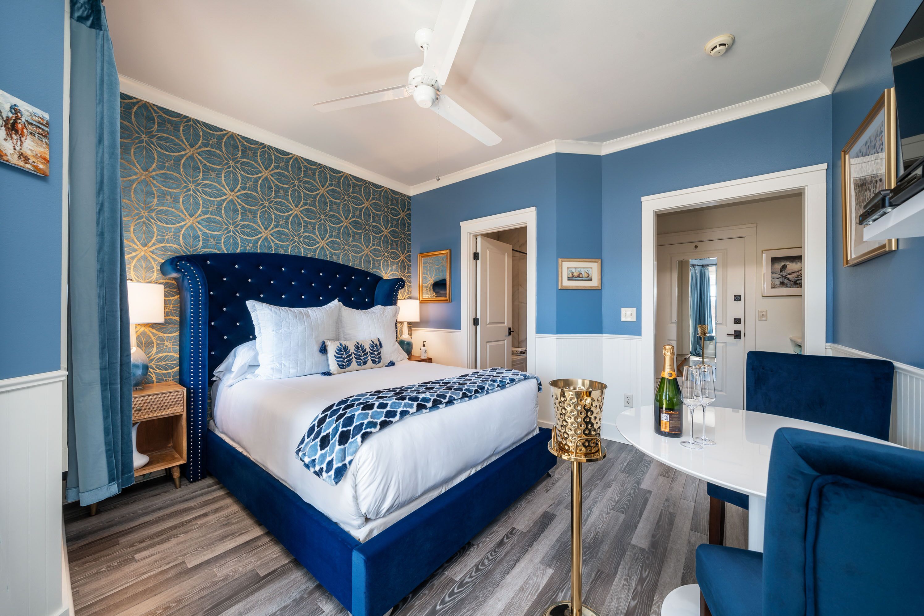 A bedroom with a large bed, a bedside table, a nightstand, a ceiling fan, a small table with a bottle and glasses, and a chair.