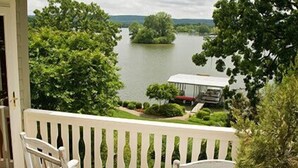 Un balcon avec des chaises blanches et une table, donnant sur un lac avec un quai et une maison en arrière-plan.