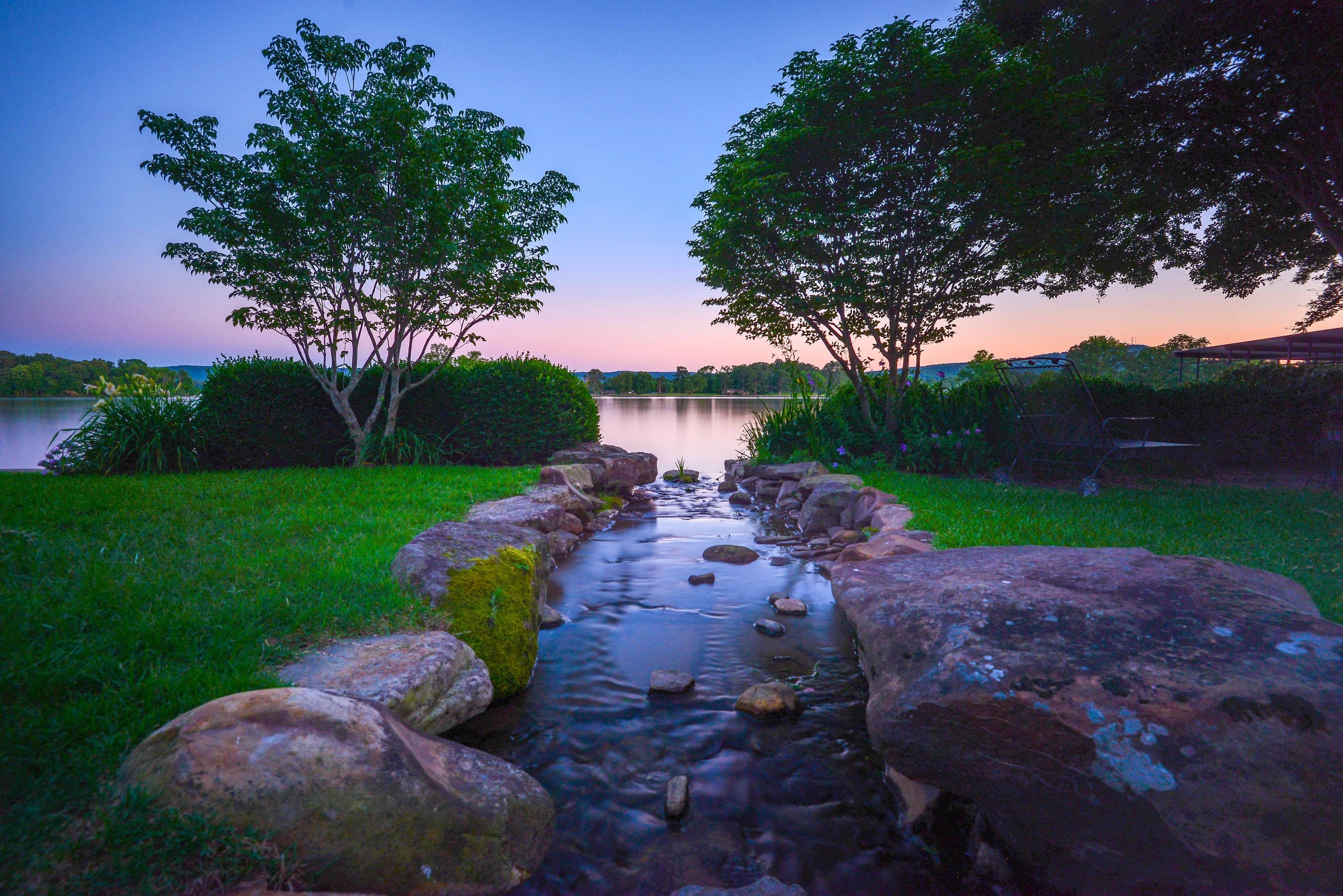 A serene outdoor scene with a stream flowing through a rocky bed, surrounded by lush greenery and trees.