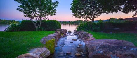 A serene outdoor scene with a stream flowing through a rocky bed, surrounded by lush greenery and trees.