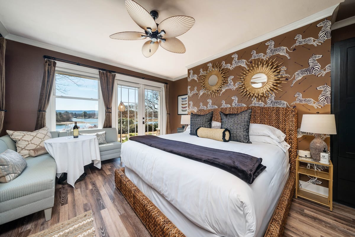 A bedroom with a large bed, a ceiling fan, a wall-mounted mirror, and a view of the outdoors.