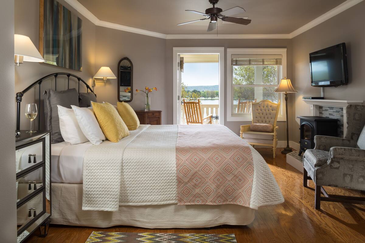 A bedroom with a bed, bedside lamps, a TV, a fireplace, and a view of the outdoors.