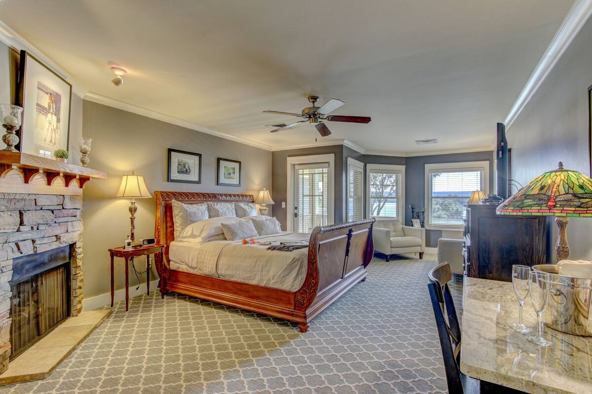 A bedroom with a large bed, a fireplace, a ceiling fan, and a dining area with a table and chairs.