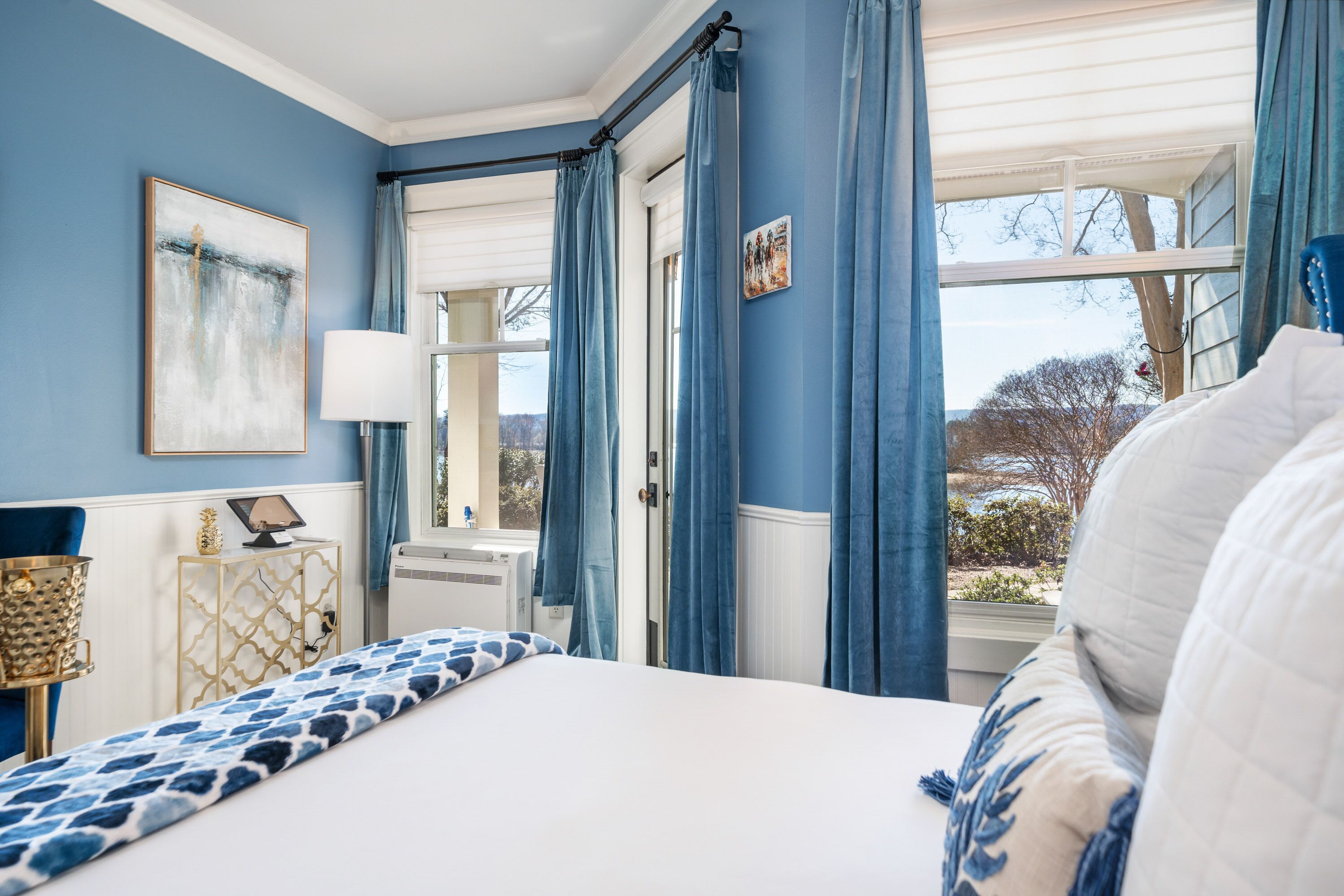 A bedroom with a bed, a bedside table, a lamp, a window with curtains, and a view of the outdoors.