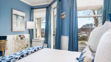A bedroom with a bed, a bedside table, a lamp, a window with curtains, and a view of the outdoors.