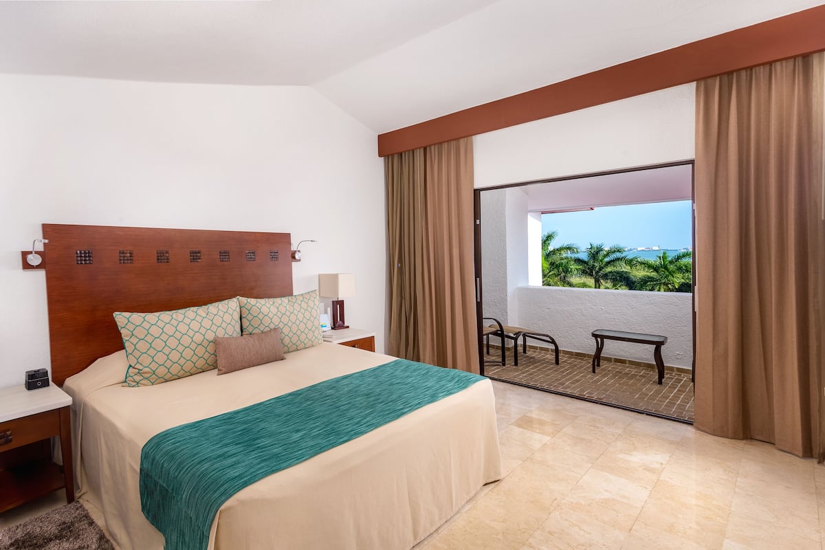 Habitación de hotel con cama, mesitas de noche, un balcón con vistas y una ventana con cortinas.