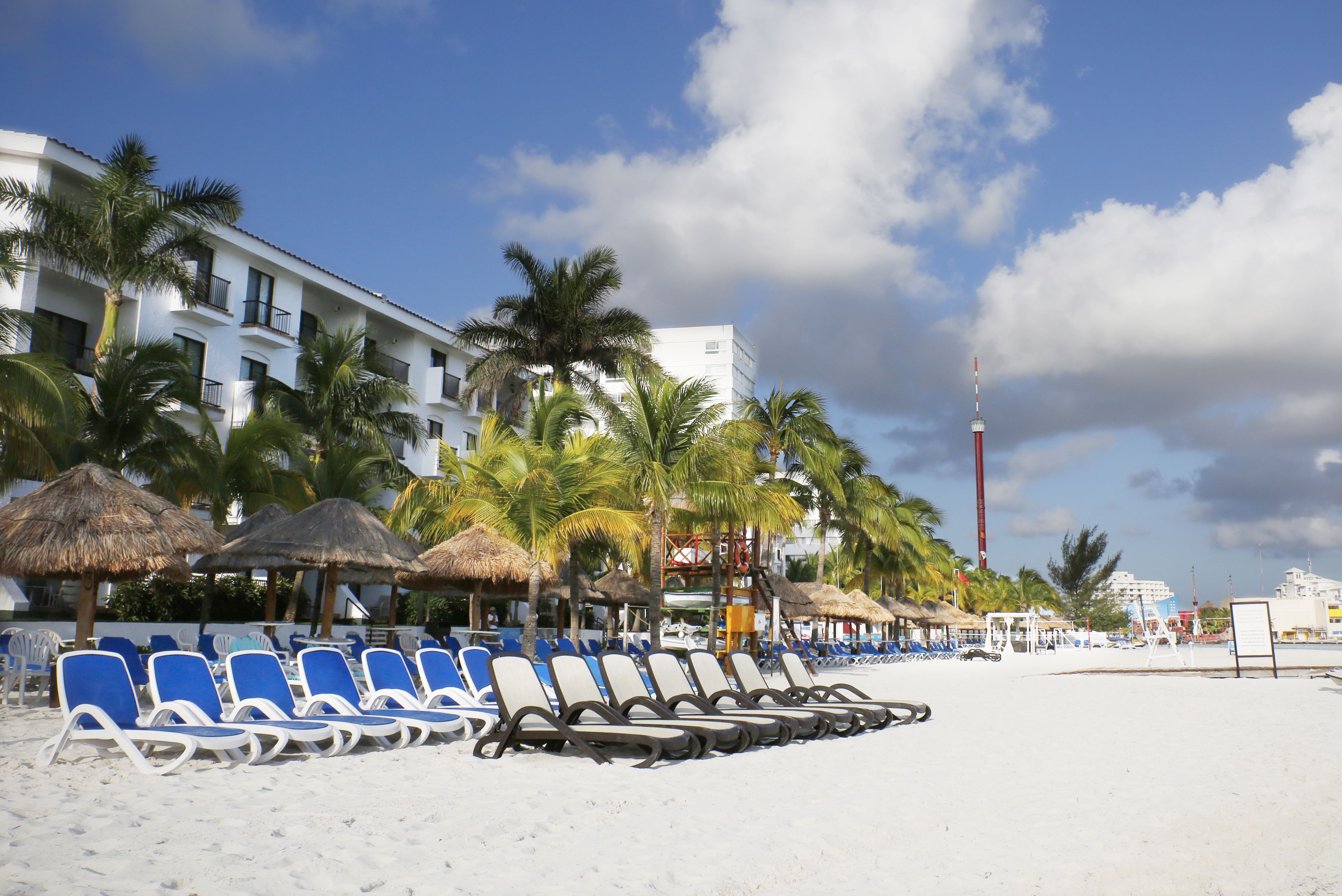A beach with rows of blue lounge chairs, palm trees, and a hotel building with balconies.