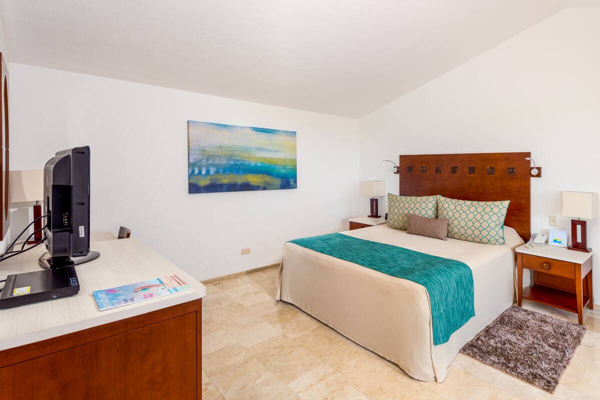 Habitación de hotel con cama, escritorio con computadora, televisor y un cuadro en la pared.