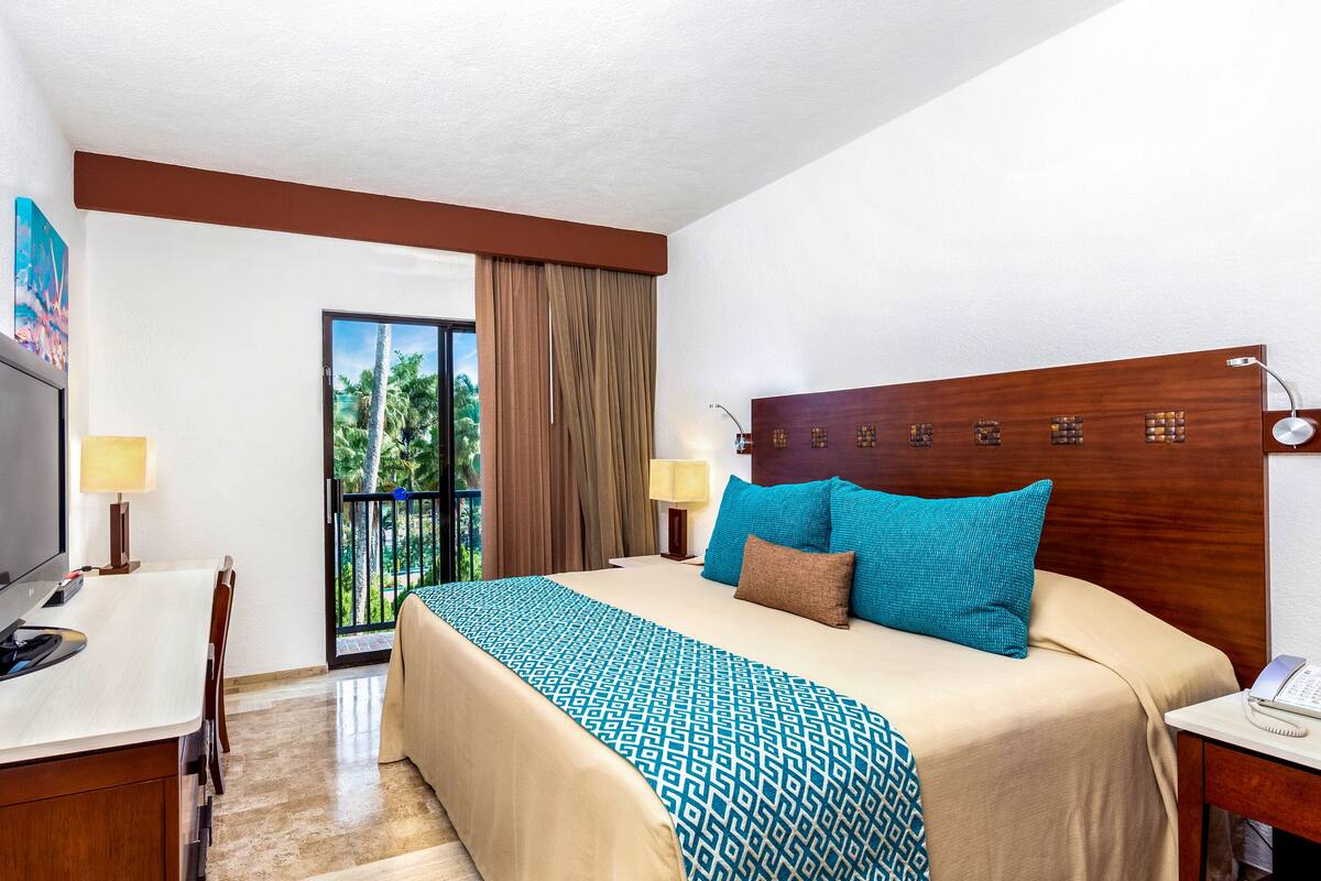 Habitación de hotel con cama, escritorio, computadora, teléfono y ventana con vista a palmeras.